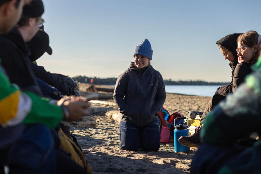 A person wearing winter clothing kneels on a sandy beach in the winter sun with a city and ocean in the background while a group of people sit on beach logs to the left and right.