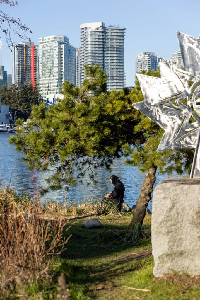 A person in winter clothes sits on a sunny day in the middle distance on a semi-wild space framed by pine trees and sculptures at the edge of a waterway against a backdrop of glass-fronted towers.