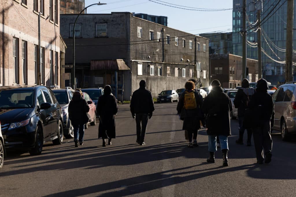 A group of people with backs turned walk in the winter sun down a road in an industrial area, their shadows long amidst parked cars reflecting the pale light.