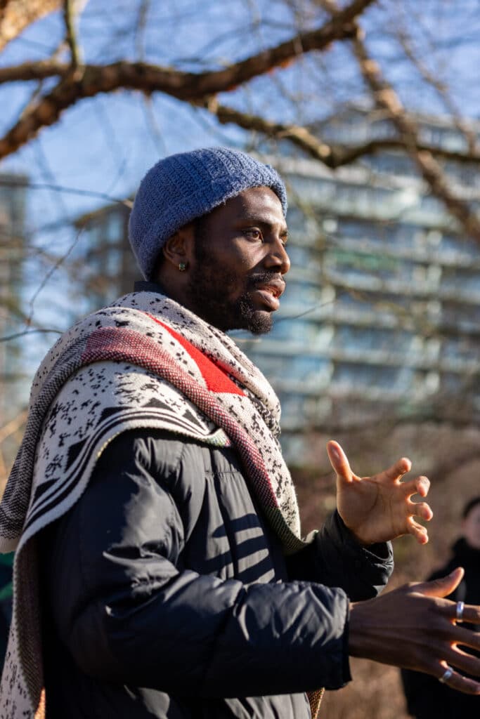 A person wearing winter clothing stands speaking in the winter sun against a background of leafless trees and a wide, gridded, multistory building.