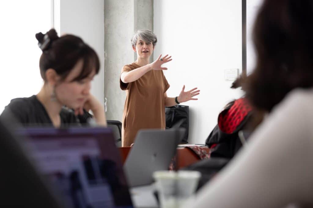 A person stands at the head of a classroom speaking to several seated students.