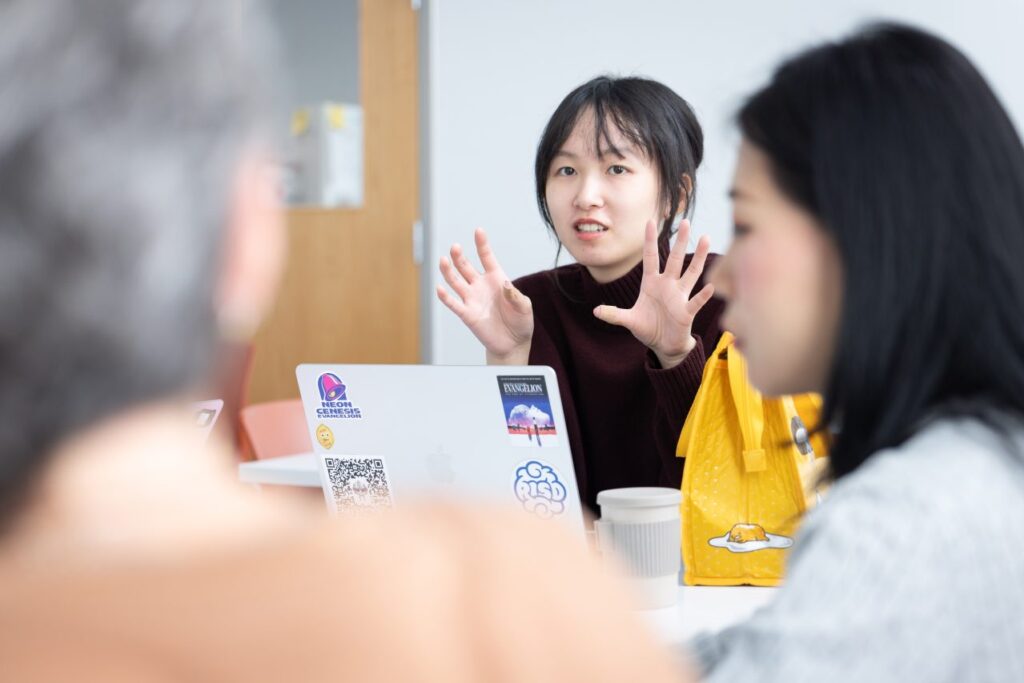 A person seated at a table with a laptop gestures with their hands while they speak to two other people partially visible in the foreground.