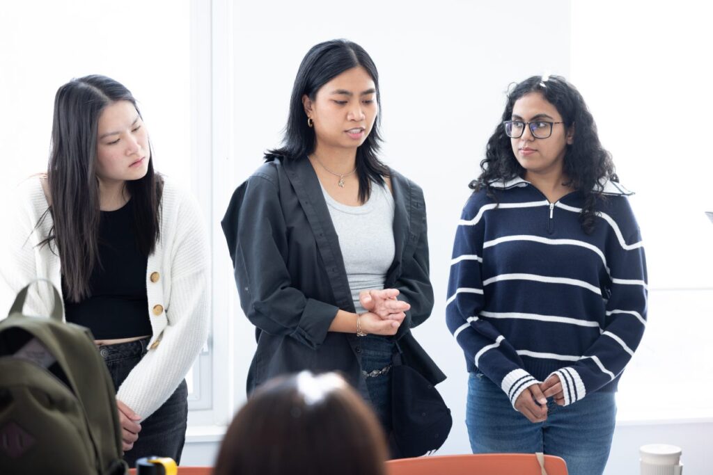 Three people stand speaking before a group of several other people who are all seated around a table.