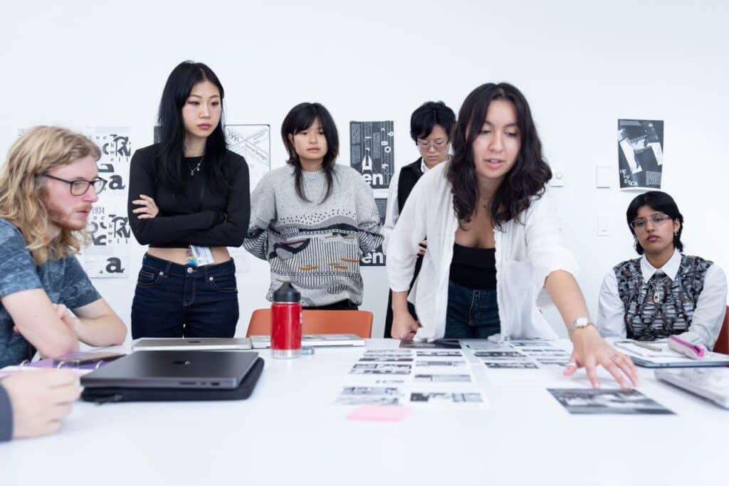 A group of people sit and stand chatting around a table on which are arranged several pieces of printed paper.