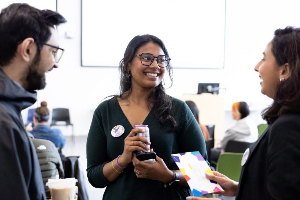 A person in centre of frame smiles broadly while speaking with two others, one to each side of the frame.