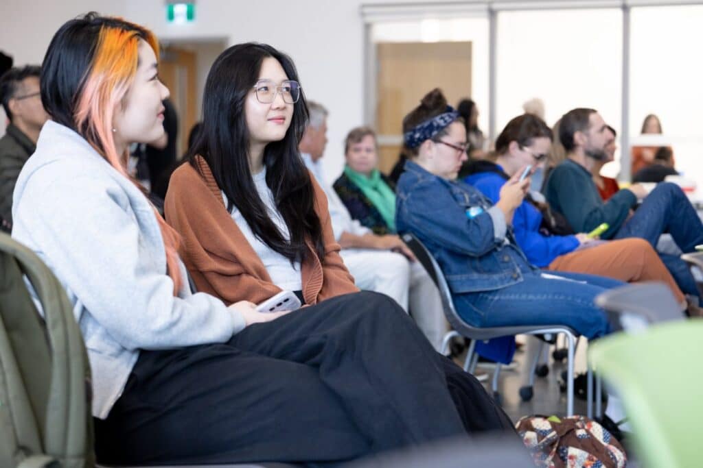 Two people in sharp focus amongst an audeince all seated in a classroom smile as they listen to a speaker.