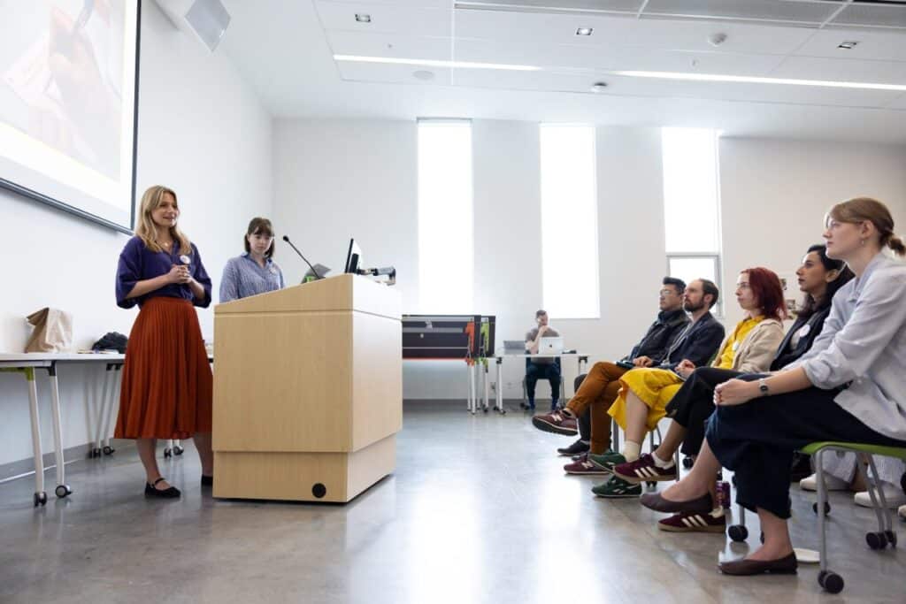 Two people stand at a lectern speaking into a microphone beneath a projection screen to a seated audience in a room with tall vertical sunlit windows.
