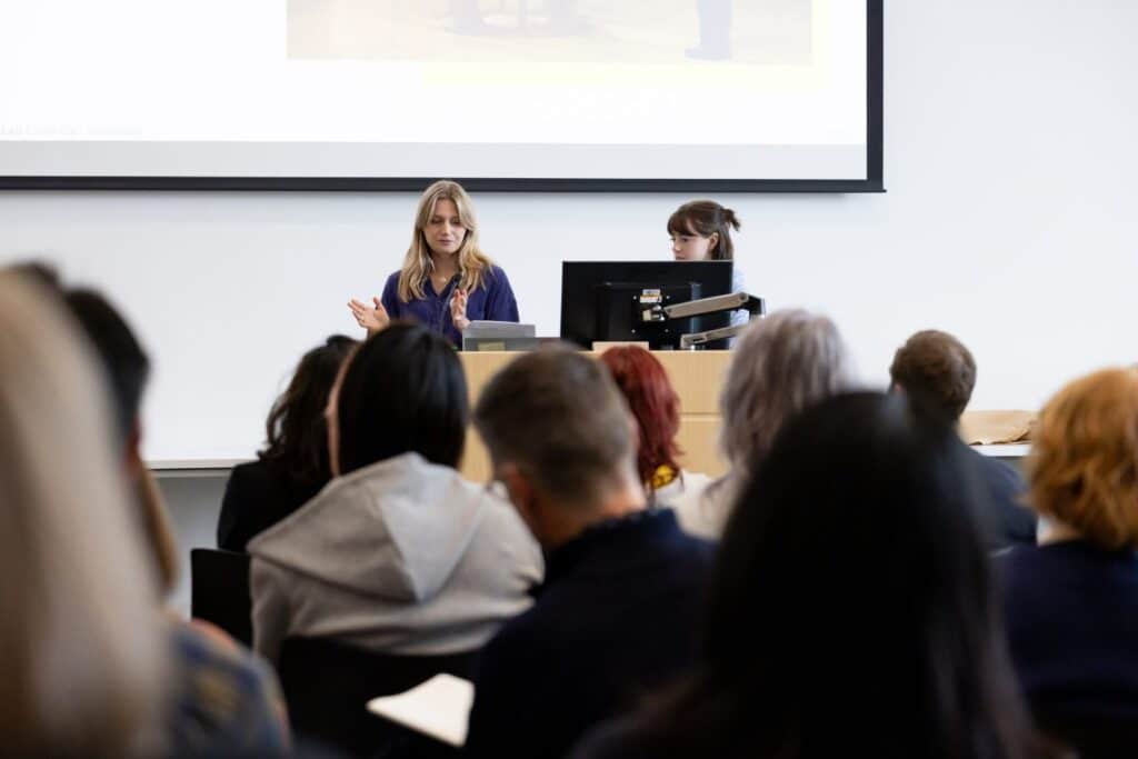 A person speaks at a podium in a classroom while another operates a computer, with an audience seated and listening in the foreground.