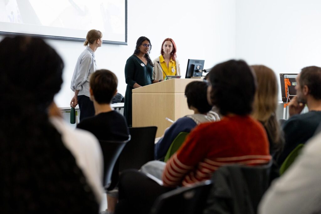 A trio of people stand at a lectern speaking into a microphone while an audience listens.