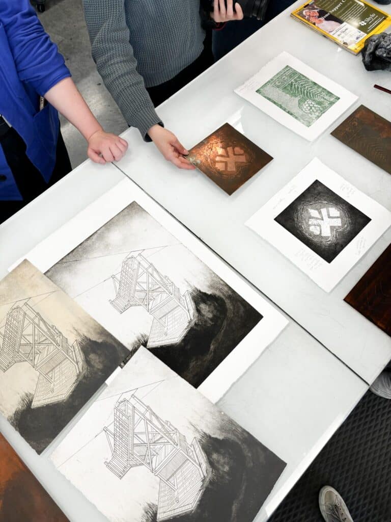 A grey table is laid with intaglio prints and copper plates, some of which are being handled by people standing mostly out of frame.