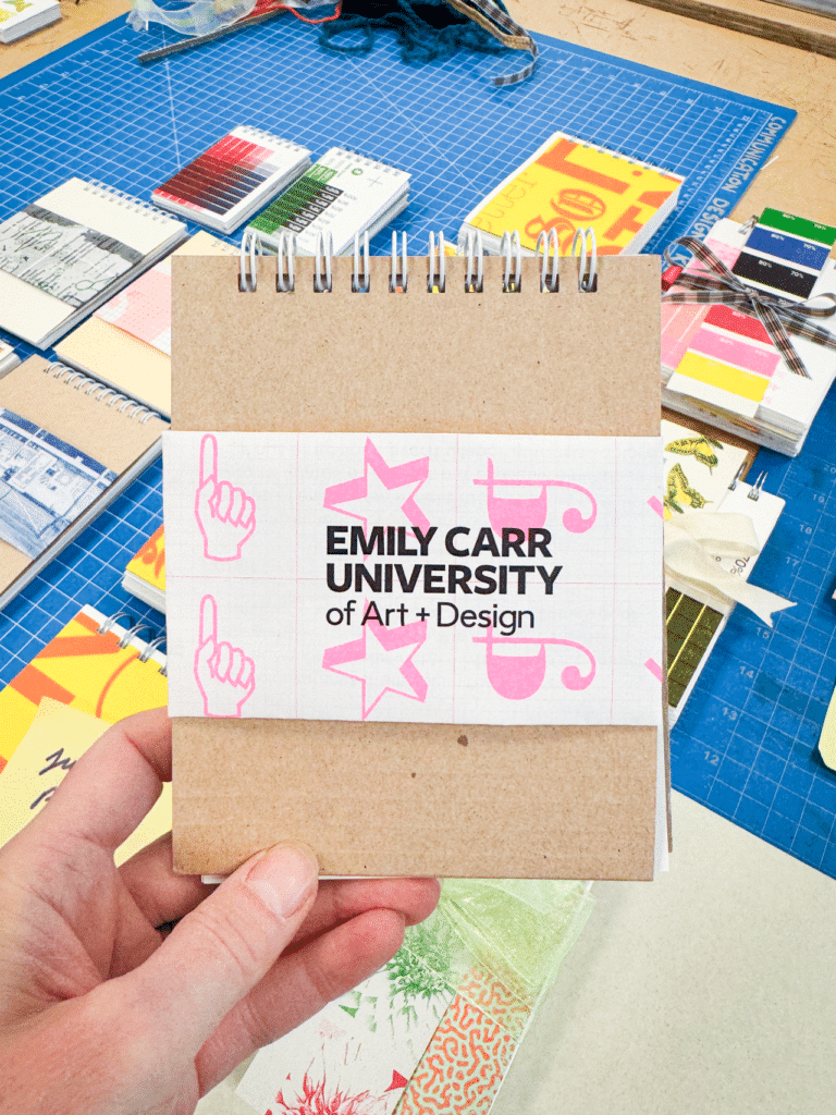 Against a backdrop of colourful stationery items arranged on a blue cutting mat, a hand holds a notepad with white spiral binding and a white belly band bearing pink graphics and black text which reads 'Emily Carr University of Art and Design.'