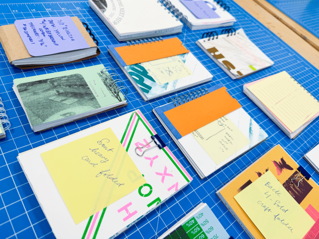 A series of spiral-bound stationery items bearing colourful graphics are arranged on a gridded blue cutting mat.