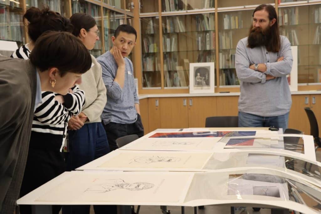 A group of people stand talking in a library around a table upon which are laid several large, colourful prints in protective plastic covers.