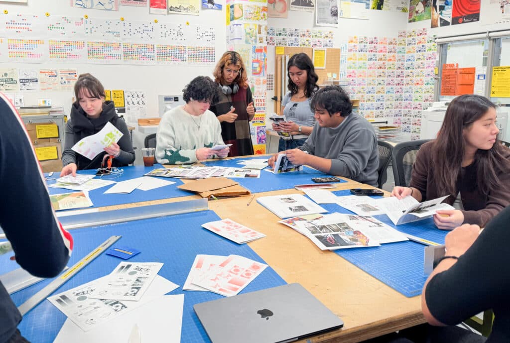 A group of people gather around a large table laid with cutting mats and printed stationery in a room whose walls are adorned with colourful printed posters.