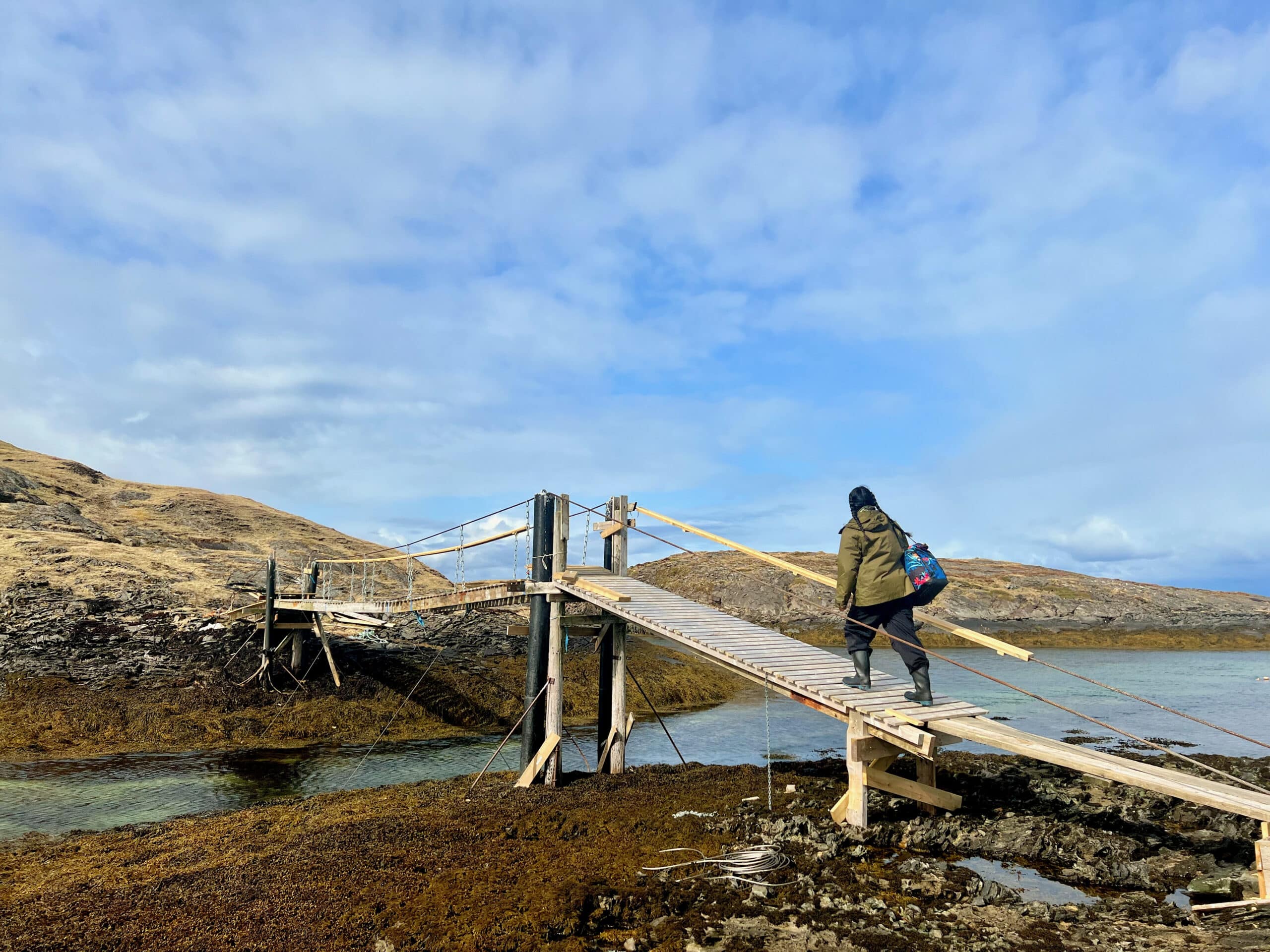 A person walking across a narrow wooden footbridge over shallow coastal water, carrying a bag, with rocky shoreline and open sky in the background.