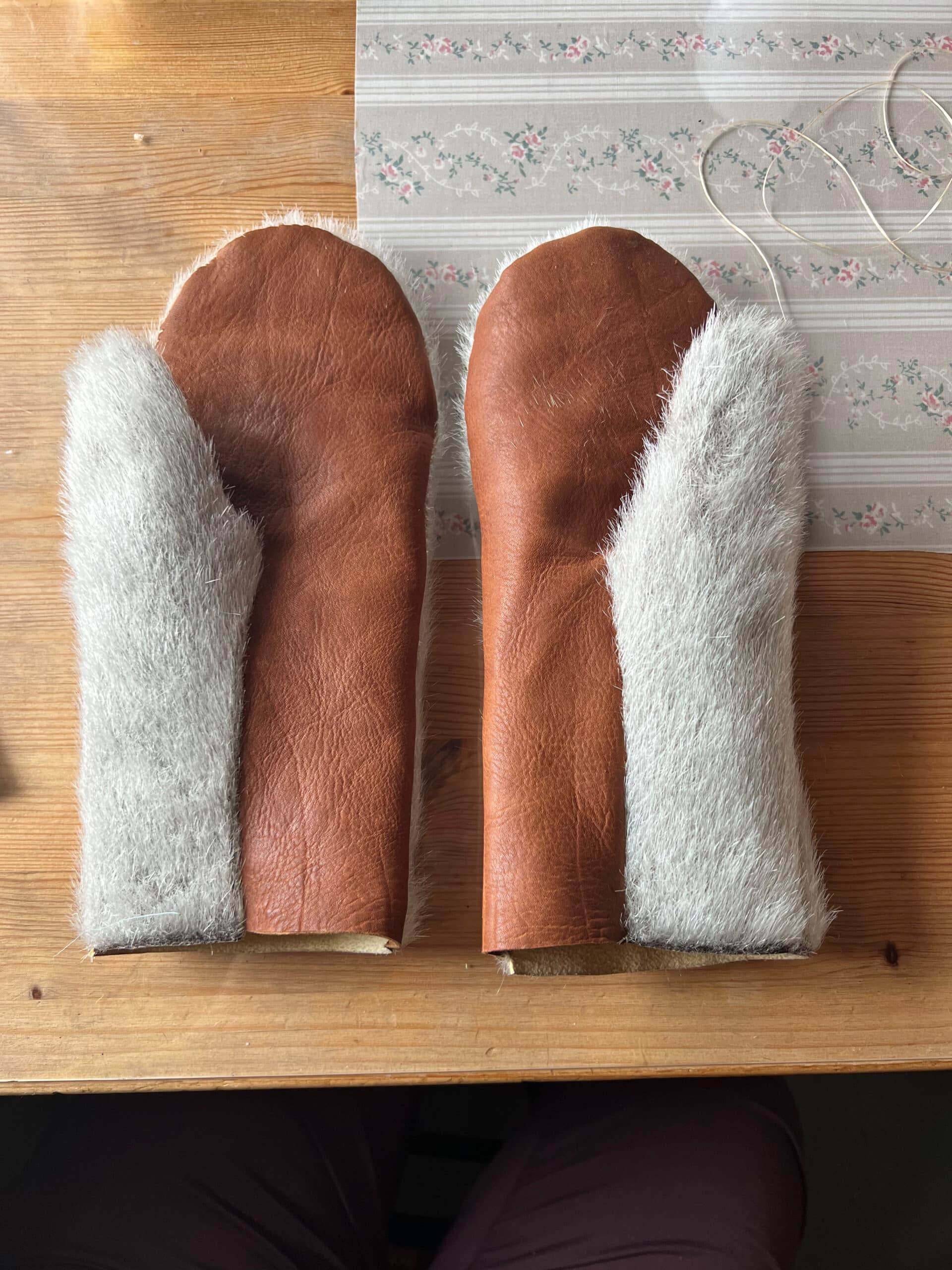 A pair of handmade leather mittens with light-coloured fur cuffs laid side by side on a wooden table.