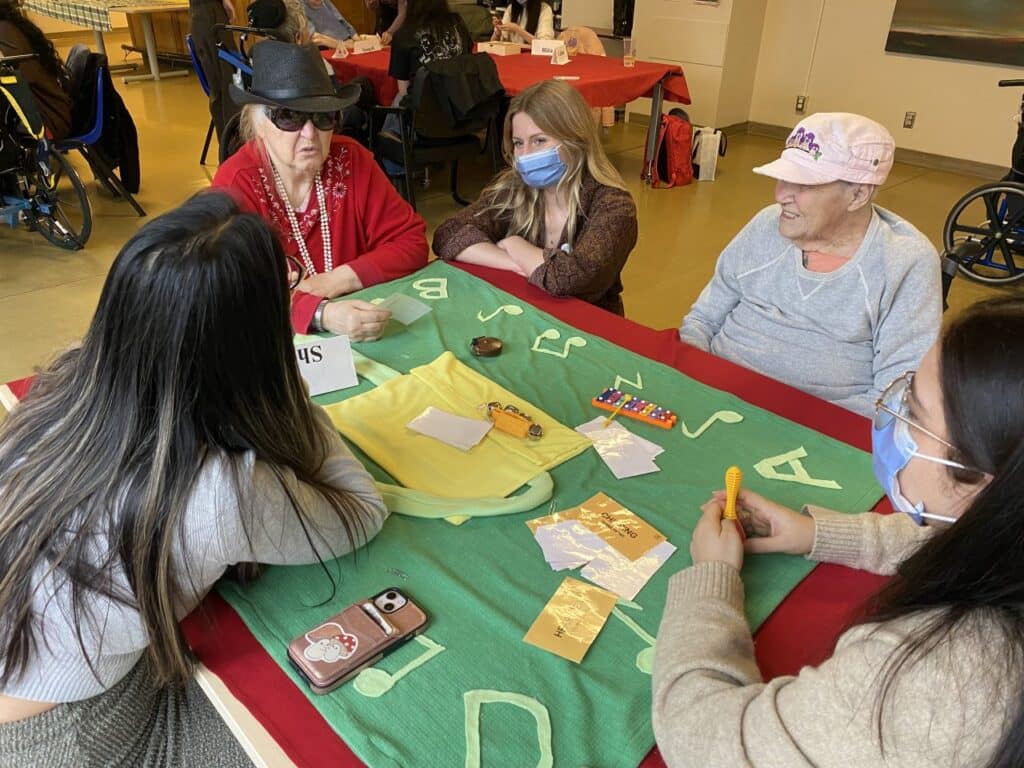 People of different ages sit around a table participating in a hands-on activity using tactile materials and cards in a communal indoor setting.