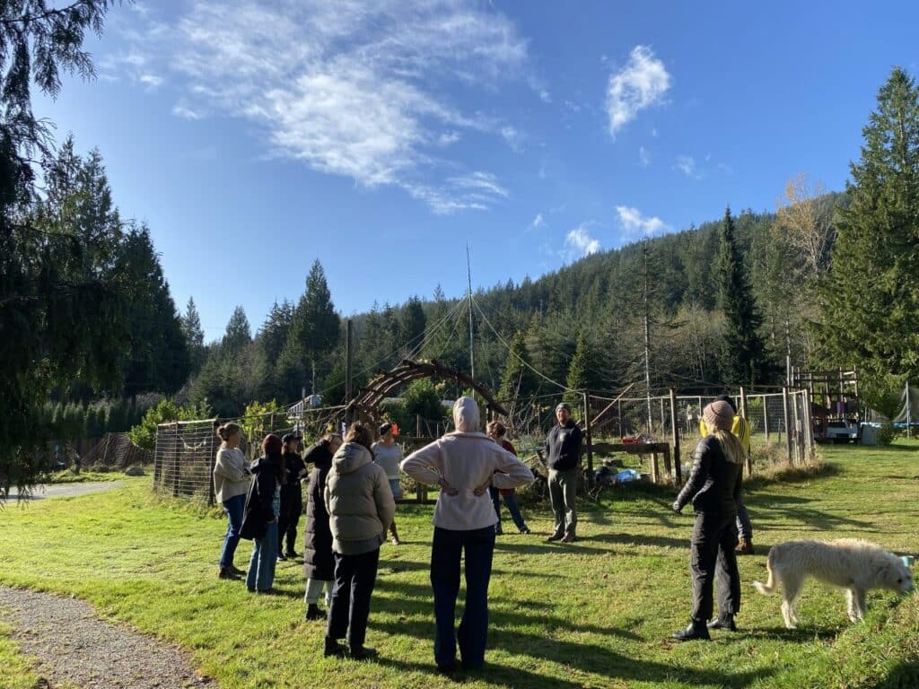 A small group stands in a grassy outdoor space surrounded by trees, listening to a speaker near a handmade structure under a clear blue sky.