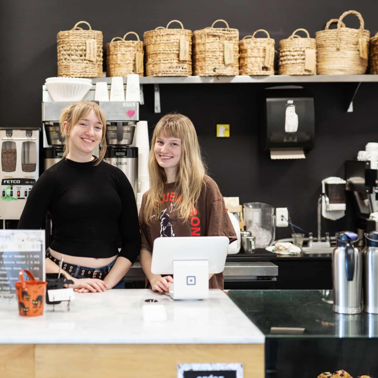 Two student baristas smile behind the counter of the campus café, The Caf.