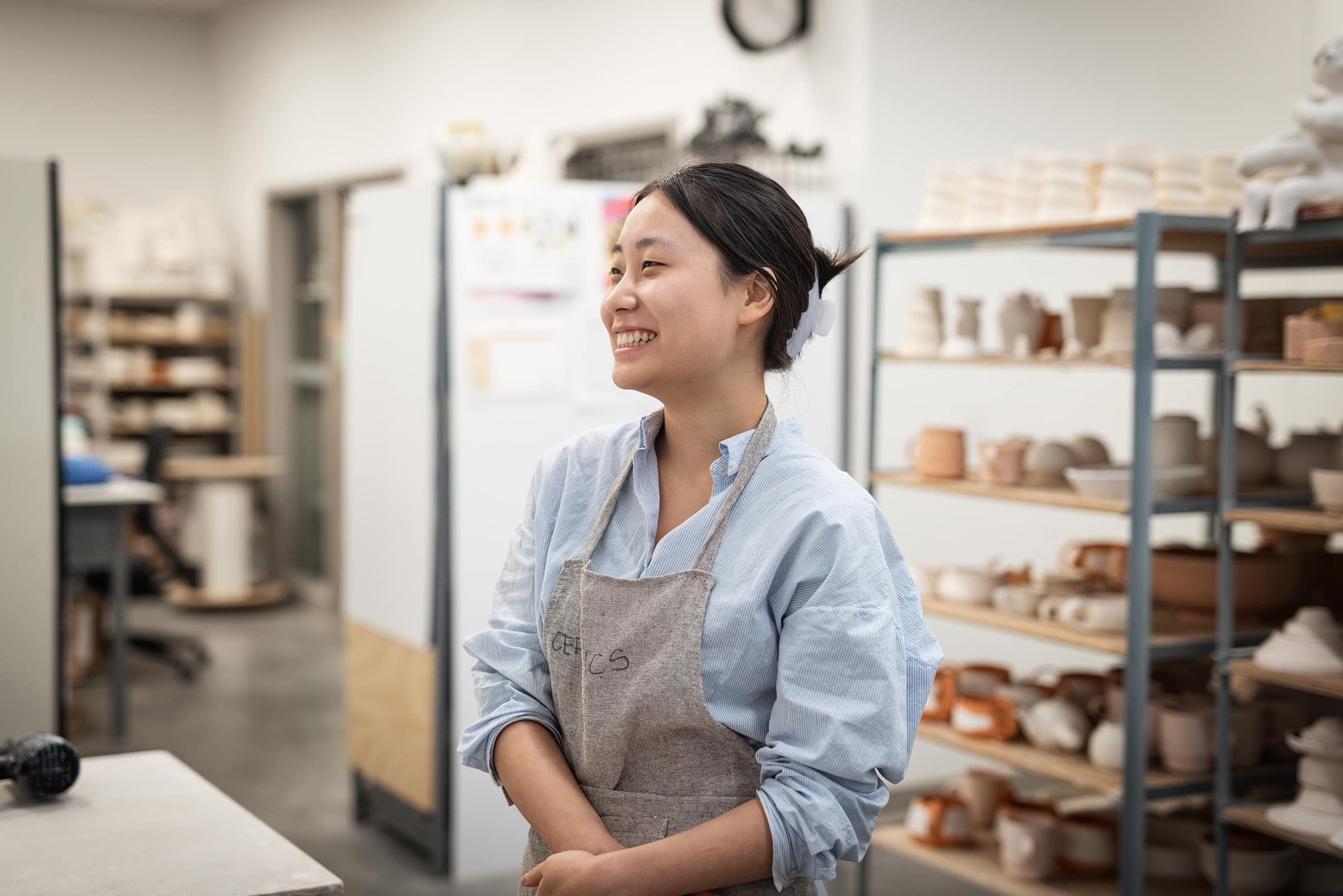 Smiling ceramic artist wearing a light blue shirt and apron stands in a pottery studio, with shelves of handmade clay pieces visible in the background.