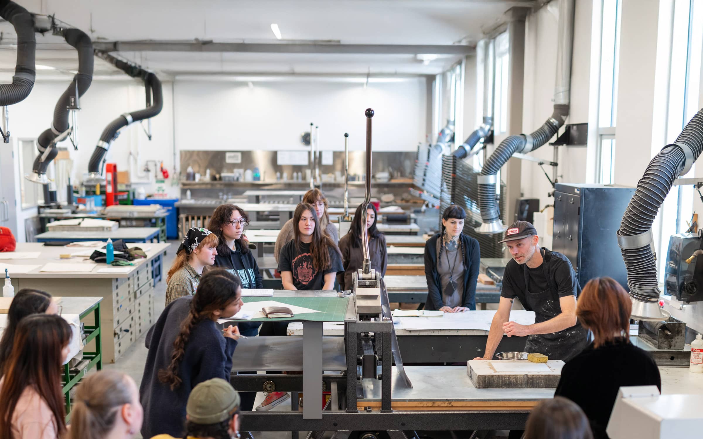 An instructor at Emily Carr University of Art + Design demonstrates traditional printmaking techniques to a group of attentive students gathered around a printing press in a well-lit, industrial-style studio with ventilation ducts overhead.