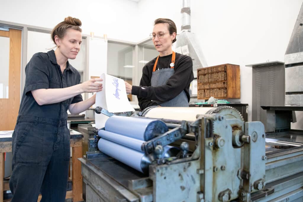 Two people wearing dark clothing work in a printmaking studio and examine a print which has been newly made on a press over which they stand.