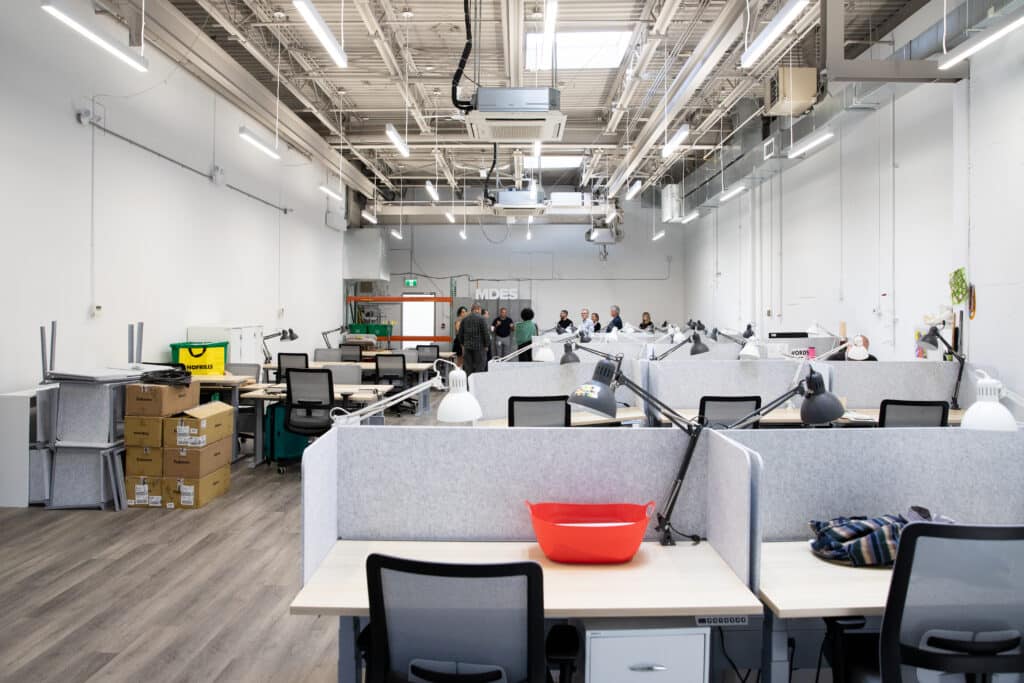A wide view of an open-plan studio with rows of desks, task lamps, and partitioned workstations, with a small group of people gathered near the back of the room.