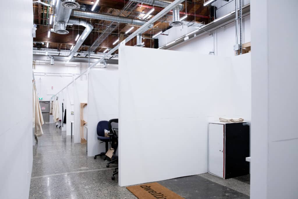 An empty corridor of a studio workspace with white partition walls, office chairs, storage cabinets, and overhead industrial lighting.