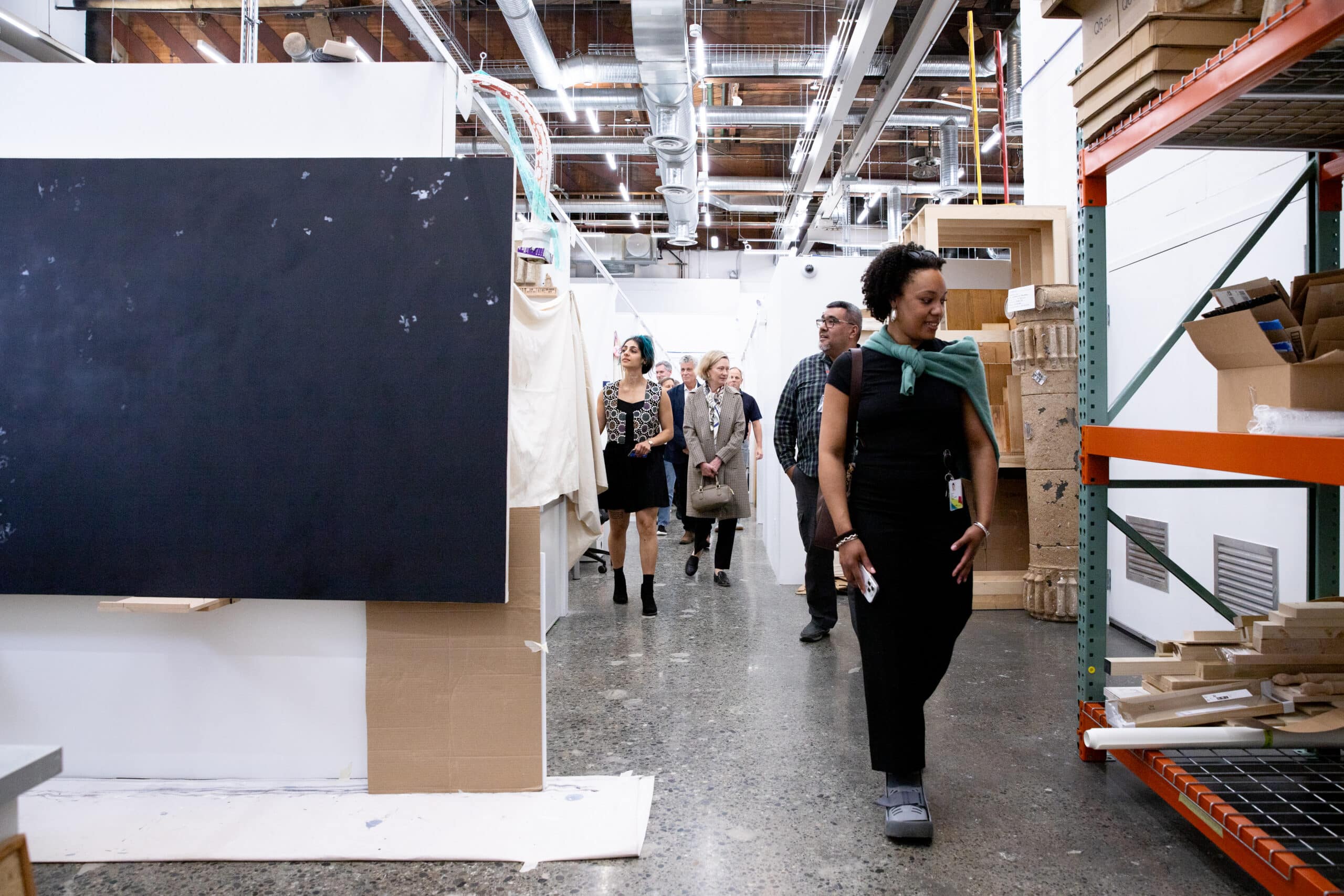 A group of people walking through a large studio workspace with high ceilings, exposed ductwork, shelving, and stacked materials, passing a large dark panel on display.