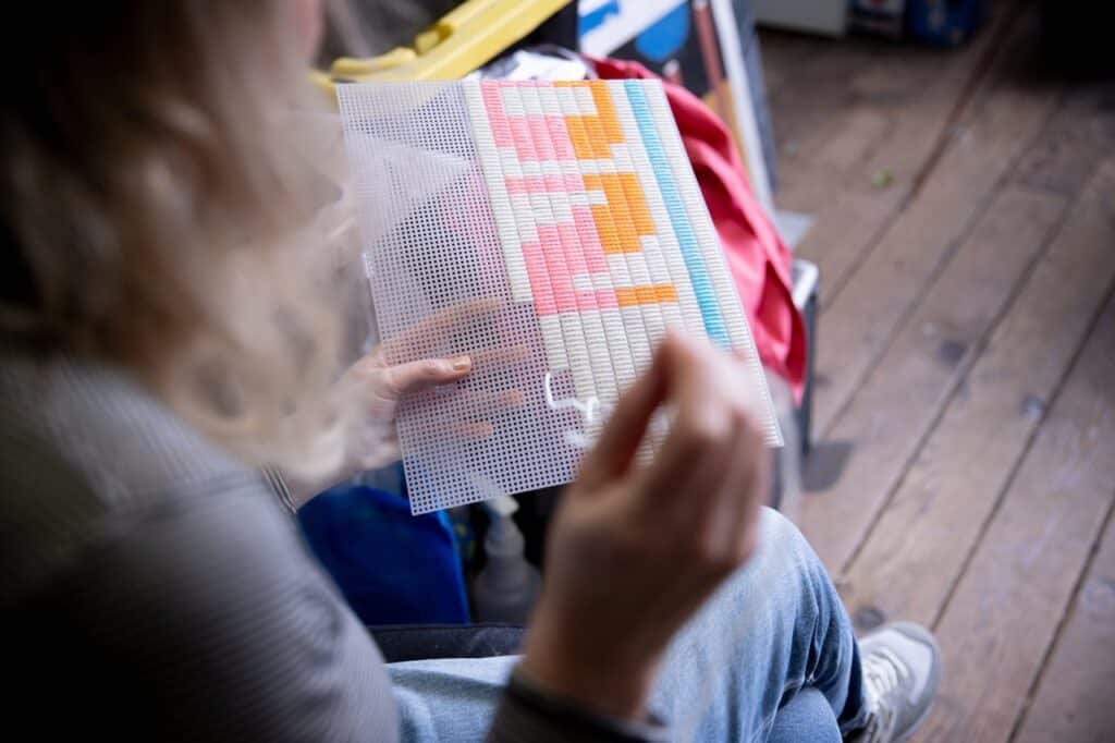 From over a person's shoulder can be seen a plastic grid held on the person's lap into which they are embroidering colourful thread in white, pink, orange and blue.