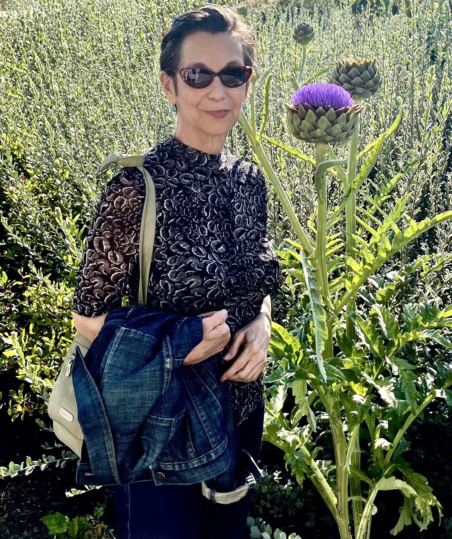 A person stands outdoors in a sunlit field beside a tall flowering thistle, wearing sunglasses and holding a jacket, with dense green plants in the background.