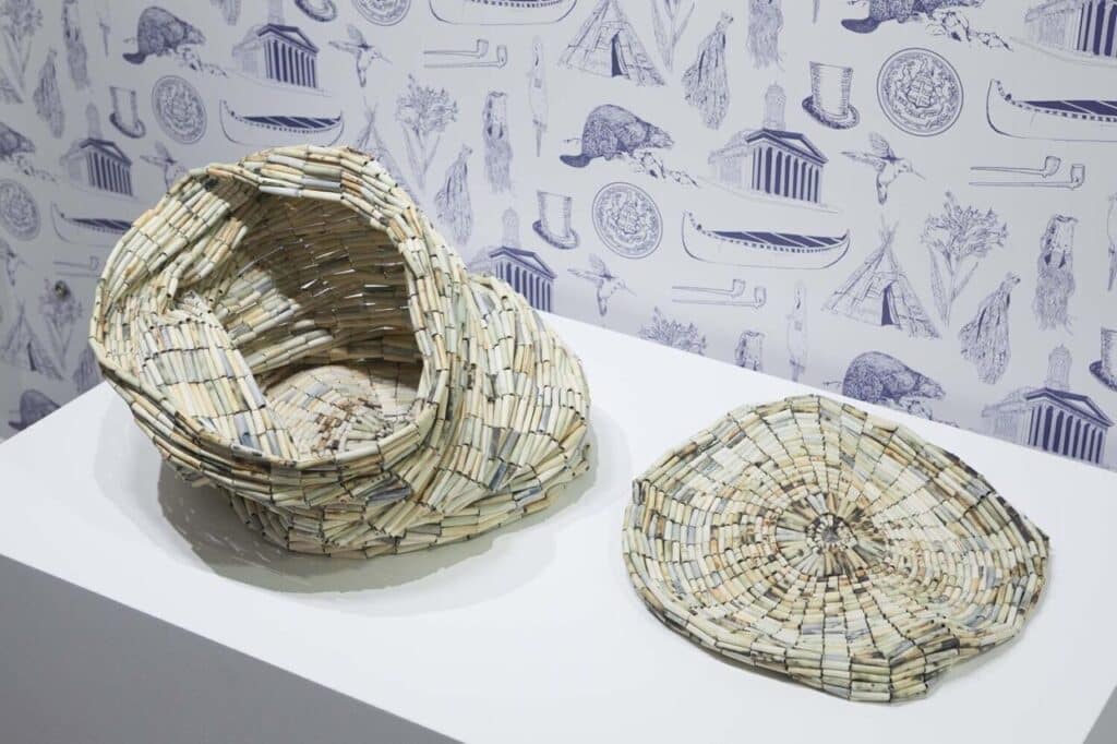 Two woven sculptural forms made from rolled paper displayed on a white plinth.