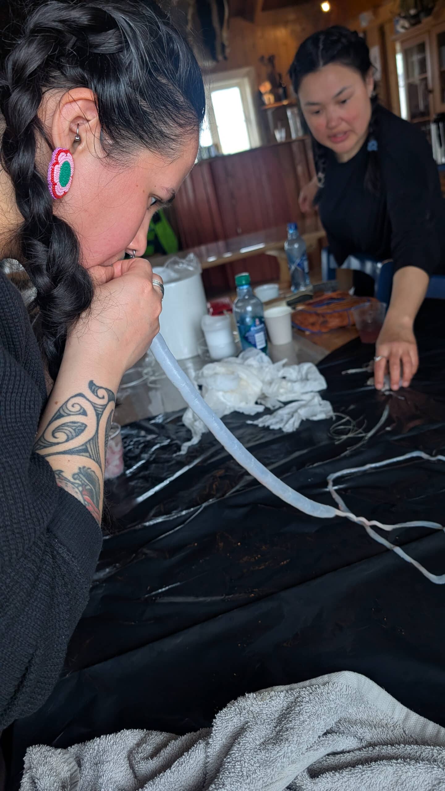 A person blowing air through a long translucent tube over a work surface covered in materials, while another person observes nearby in an indoor workspace.