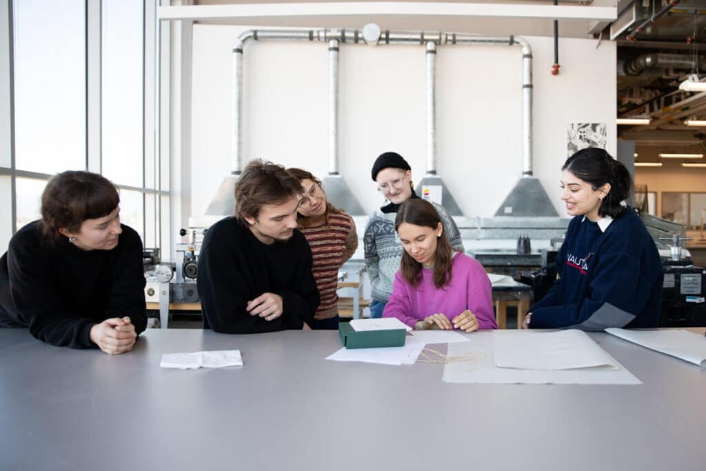 A group of people in a printmaking studio smile while they look at an unbound sketchbook spread across a wide grey desk.