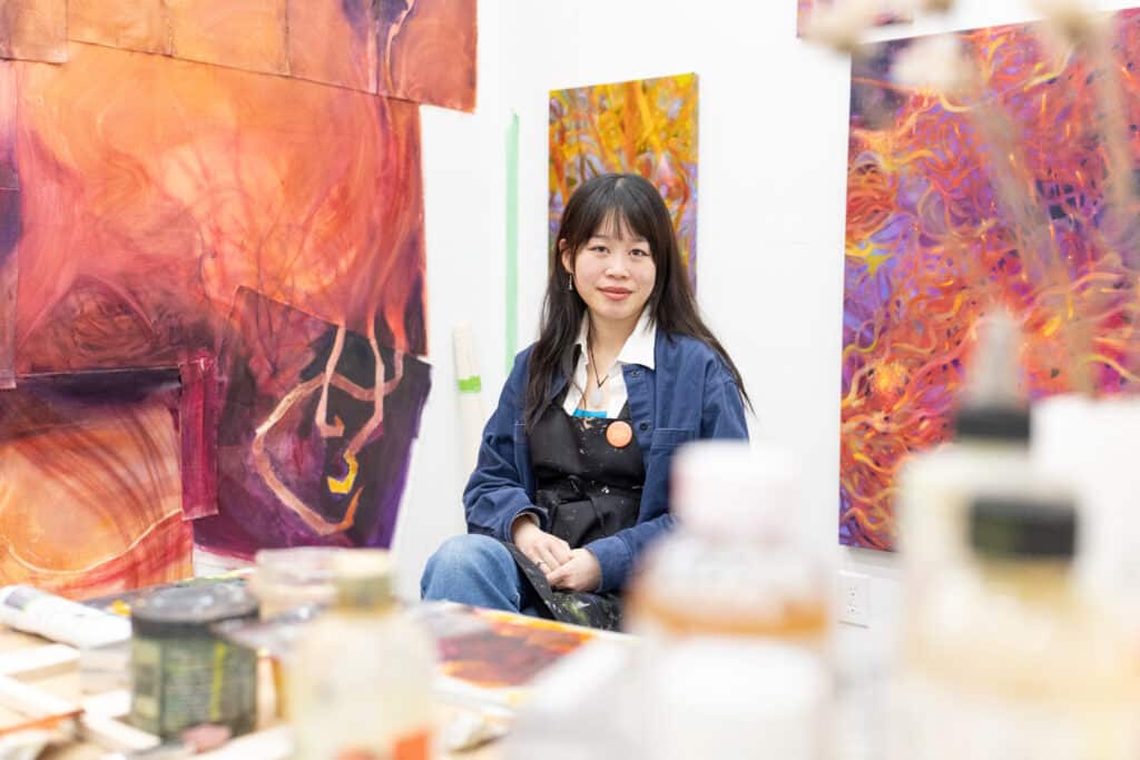 Person seated in a studio, facing the camera. They are framed by large, colorful paintings, with jars, brushes and art materials softly blurred in the foreground.