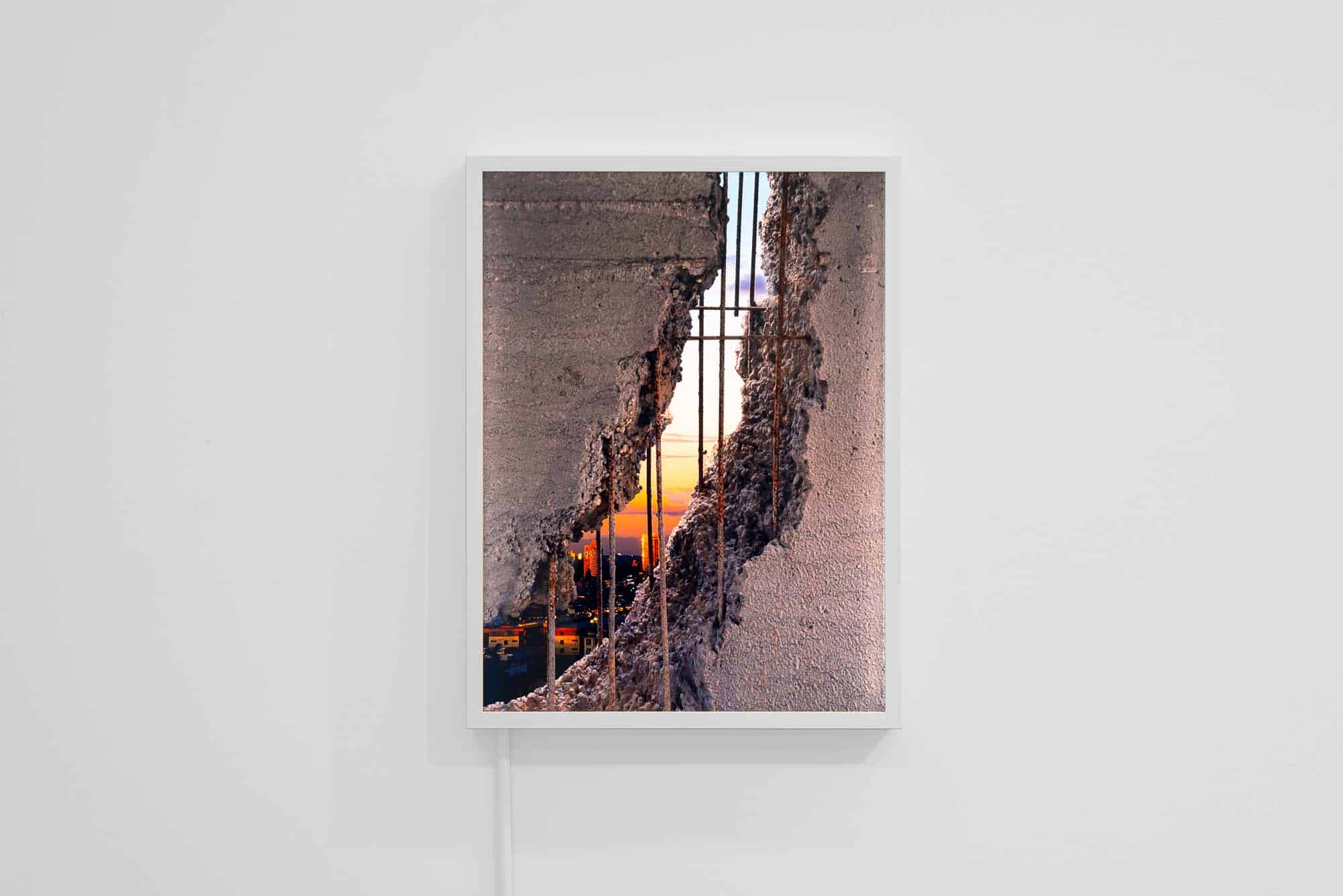 Framed photograph showing a fractured concrete wall opening onto a city skyline at sunset, with exposed metal rebar visible through the break.
