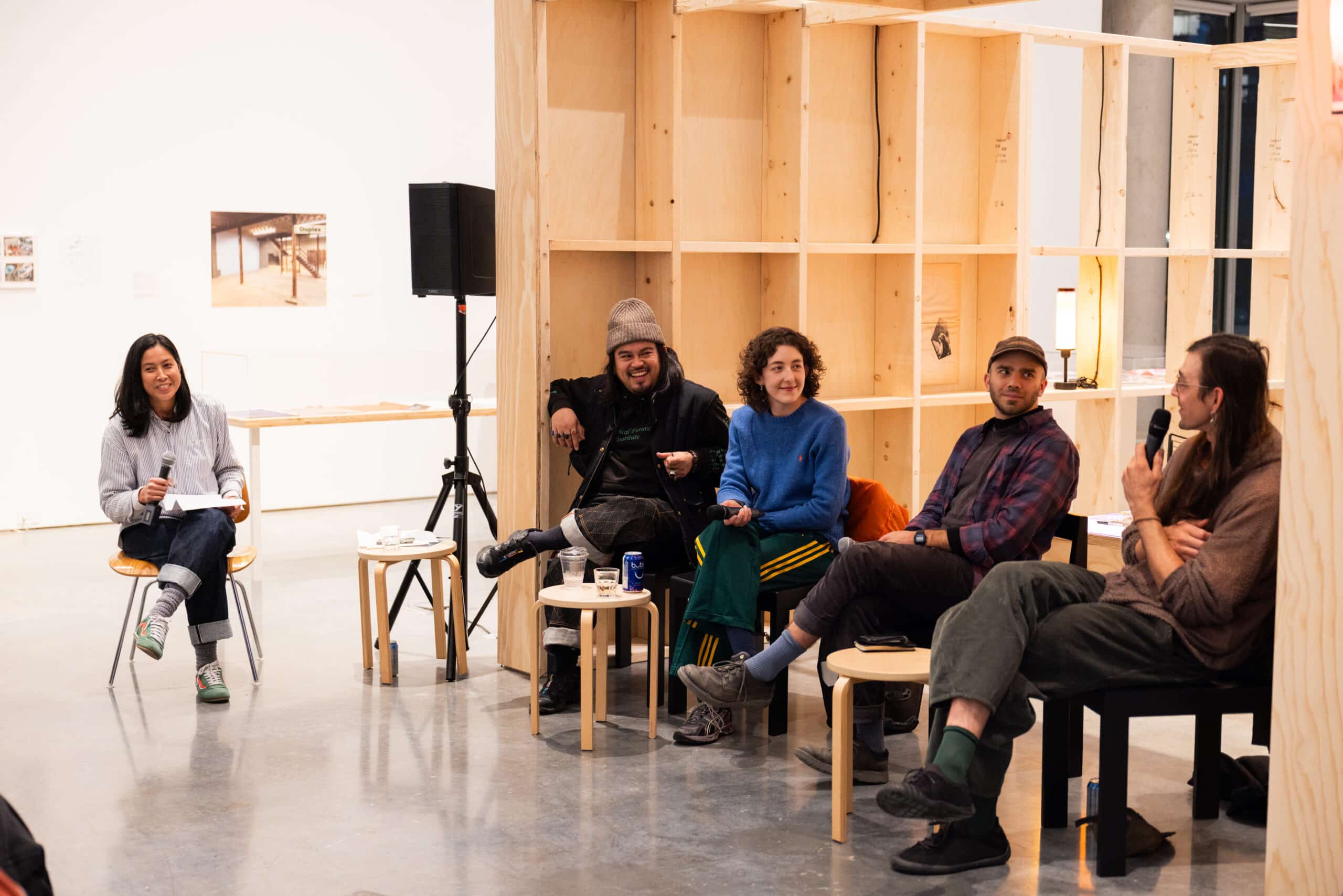 Five people seated in a panel discussion inside a gallery space, with one person holding notes on the left and four people holding microphones on the right, seated in front of a light wood shelving structure