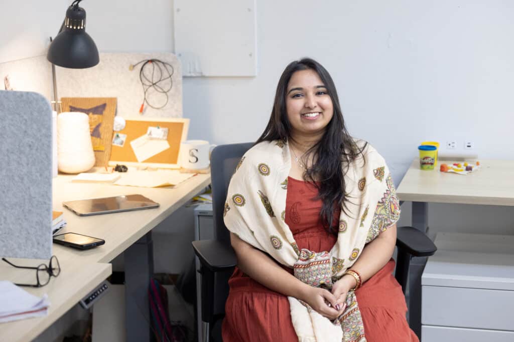 Person seated at a desk in a studio workspace, smiling toward the camera. Soft light falls across the desk, where a lamp, tablet and notes sit among personal items, with art materials and work surfaces extending into the background.