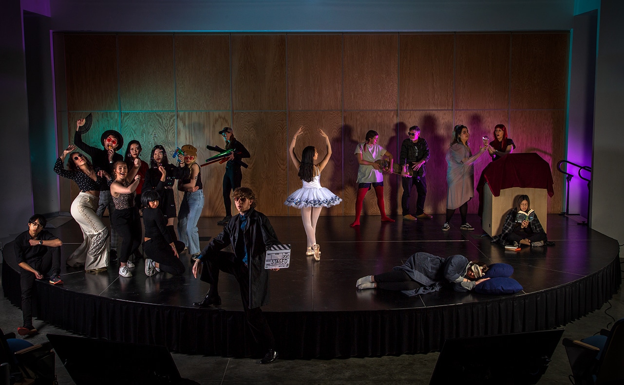 Group of performers posed on a stage in a theatrical tableau, wearing varied costumes and expressive poses under dramatic stage lighting.