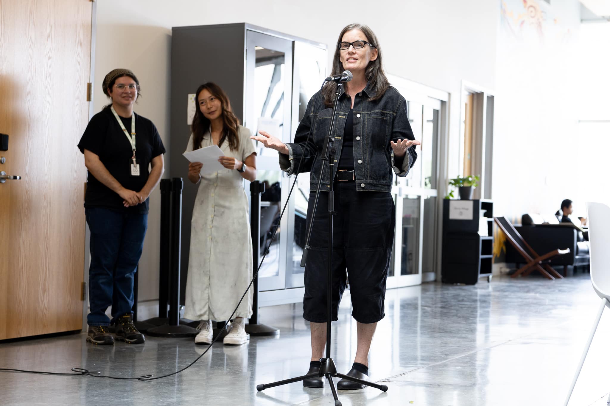 A person speaking into a microphone in an indoor gallery space, with two people standing nearby holding papers and listening.