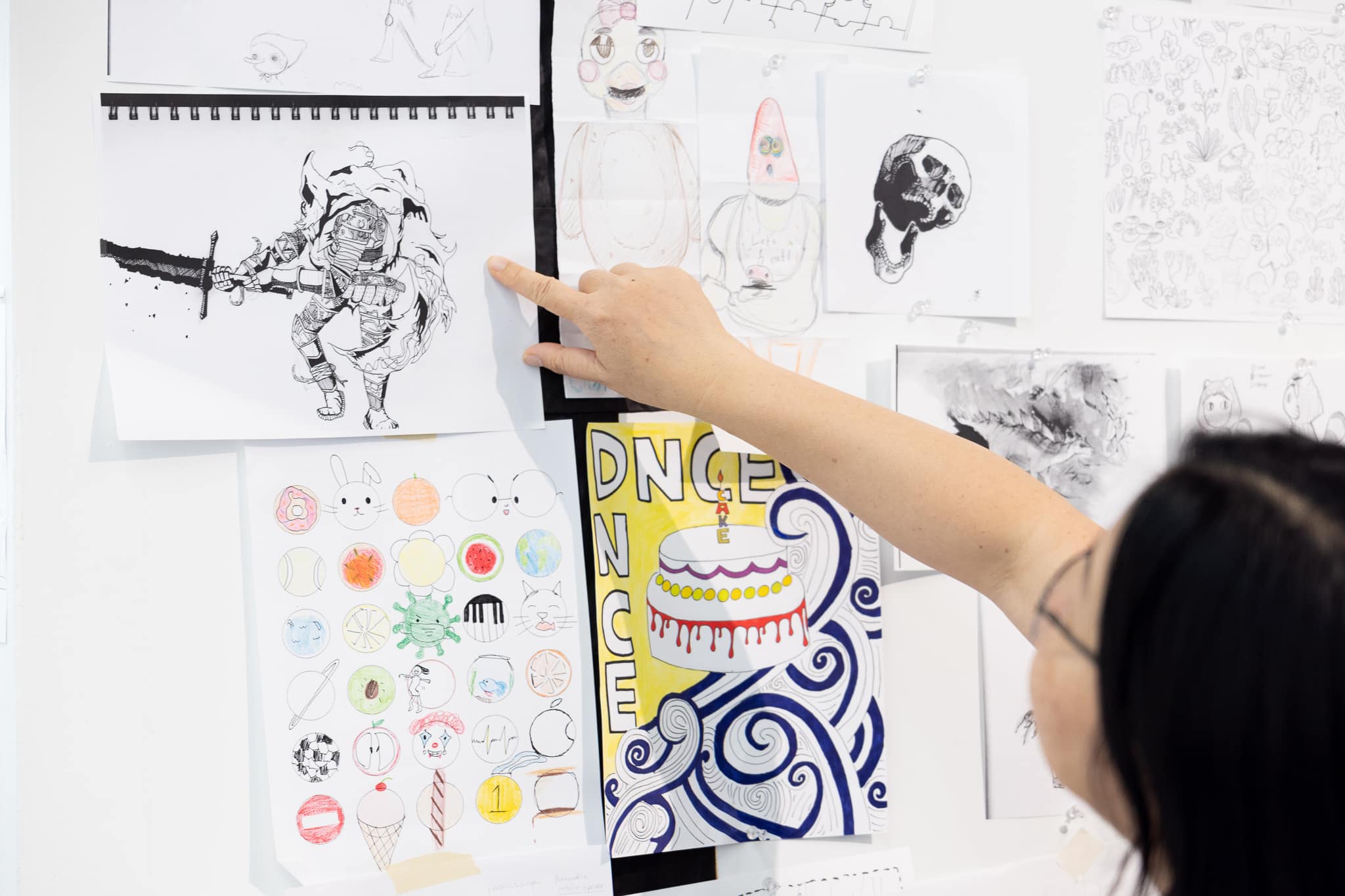 A person pointing at a wall display of student drawings and illustrations, including character sketches, patterns, and graphic designs.