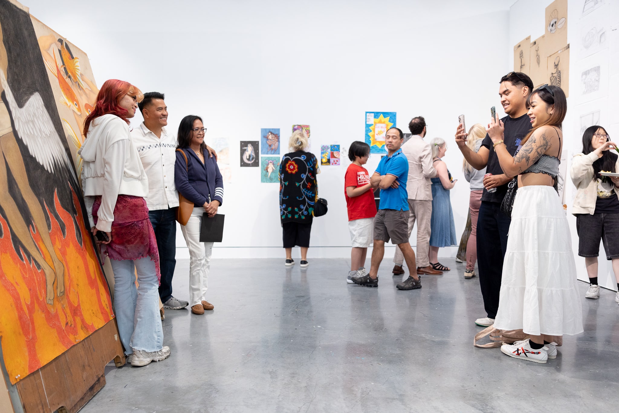 Visitors gathered in a gallery space, posing for photos and viewing artwork displayed on white walls during an exhibition.