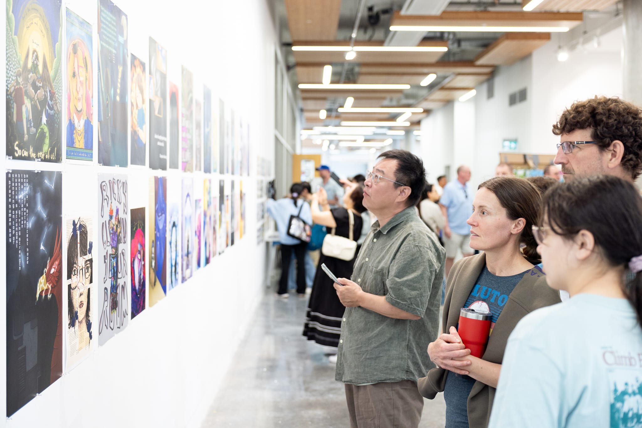 A group of people standing in a bright gallery hallway, closely viewing a wall of illustrated posters and prints arranged in rows.