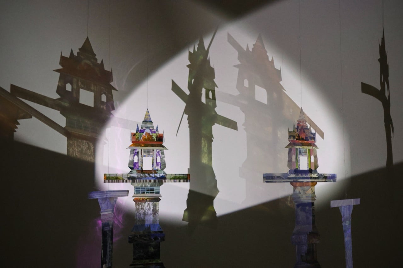 Installation featuring layered, shrine-like structures projected with patterned imagery, casting overlapping shadows onto a gallery wall.