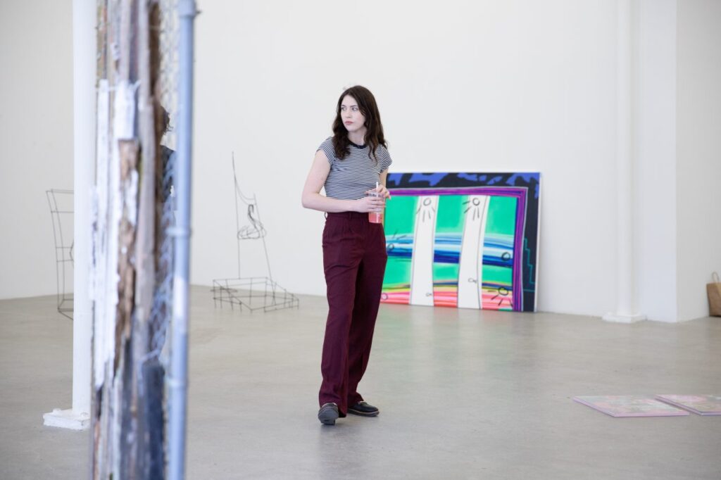 A person with lonlg hair wearing a striped t-shirt and crimson slacks looks around a gallery space filled with wireframe scupltures and colourful paintings.