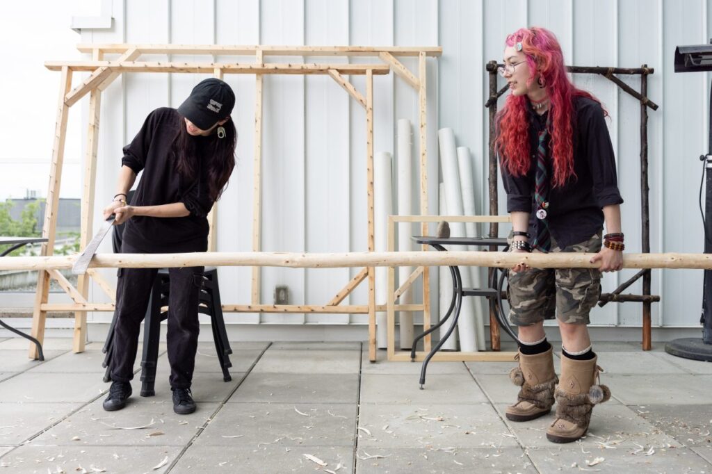 A person with pink hair and fur-lined boots holds a stripped fir pole on an outdoor patio space littered with wood shavings while a second person dressed all in black trims bark from the pole with a machete.