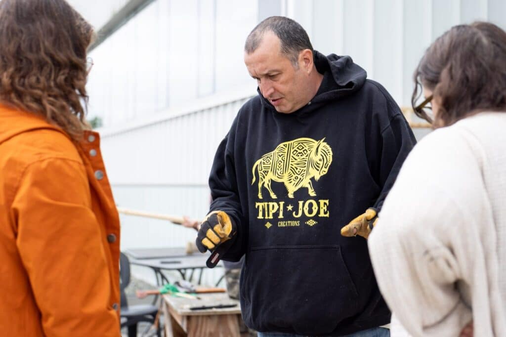 A man wearing work gloves and a black hoodie speaks with two other people on an outdoor patio space surrounded by work tools.