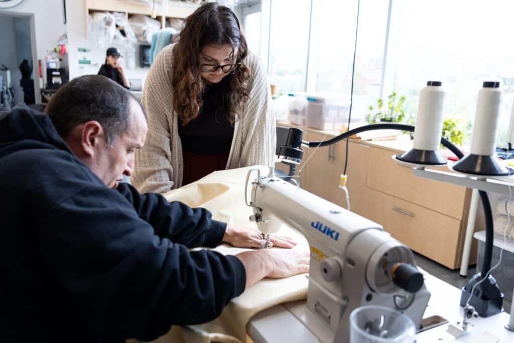 A person in a white cardigan and glasses watches another person closely as they demonstrate a technique for sewing canvas on a surger in a sunlit room.