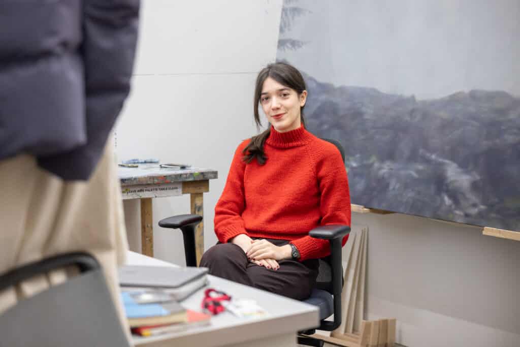 Person seated in a studio workspace, facing the camera. A red sweater stands out against worktables, art materials and a painted backdrop, creating a quiet moment within a working studio.