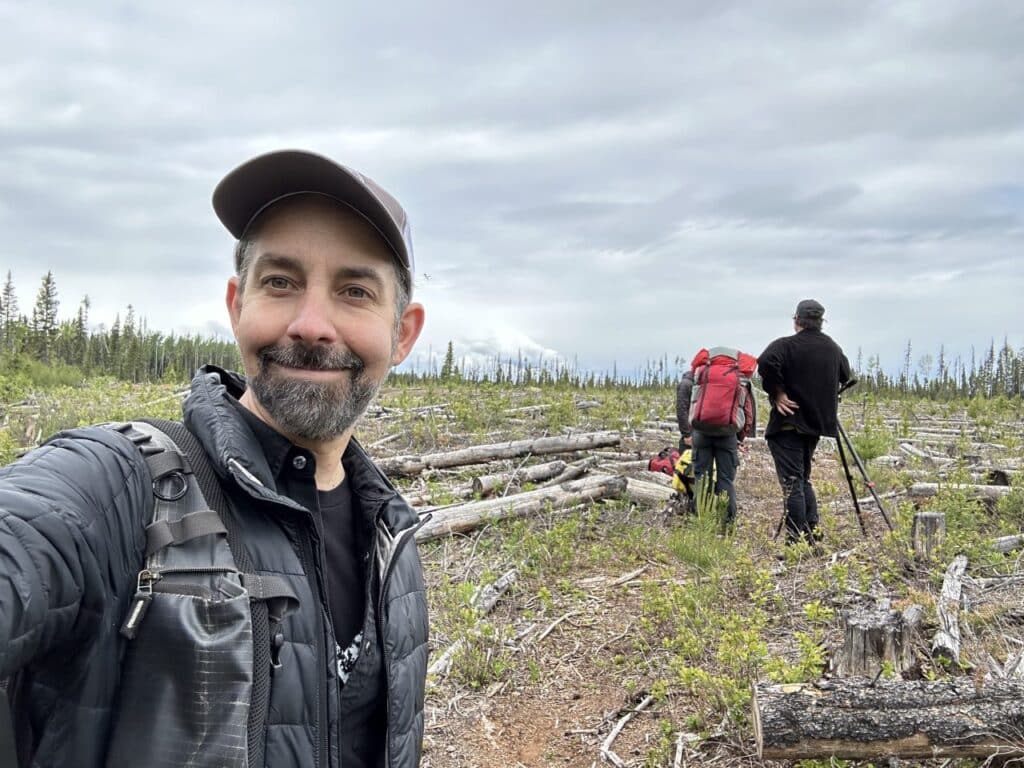 A person in a ballcap and puffer jacket smiles while standing on a scrubby mountaintop under an overcast sky while in the background a pair of people with backpacks and tripods stand amidst deadfallen wood.